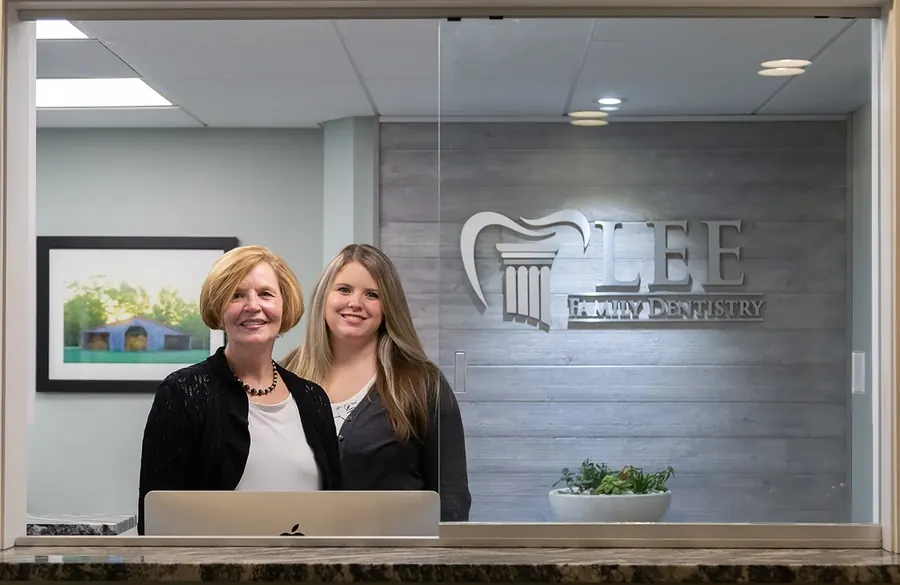 Two members of the front desk team smiling behind the reception counter, the Lee Family Dentistry sign behind them.